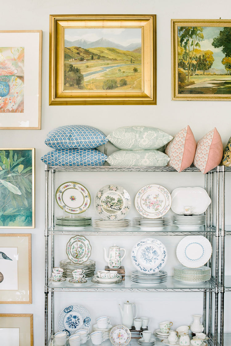 Shelves filled with vintage china, teacups, and plates, topped with pastel and patterned pillows, surrounded by framed landscape art.